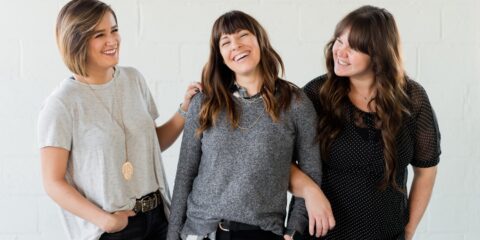 3 women smiling and standing beside white wall