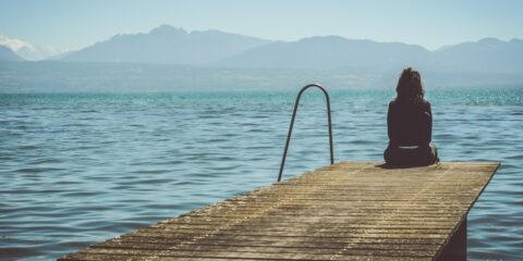 a woman sits on the end of a dock during daytime staring across a lake